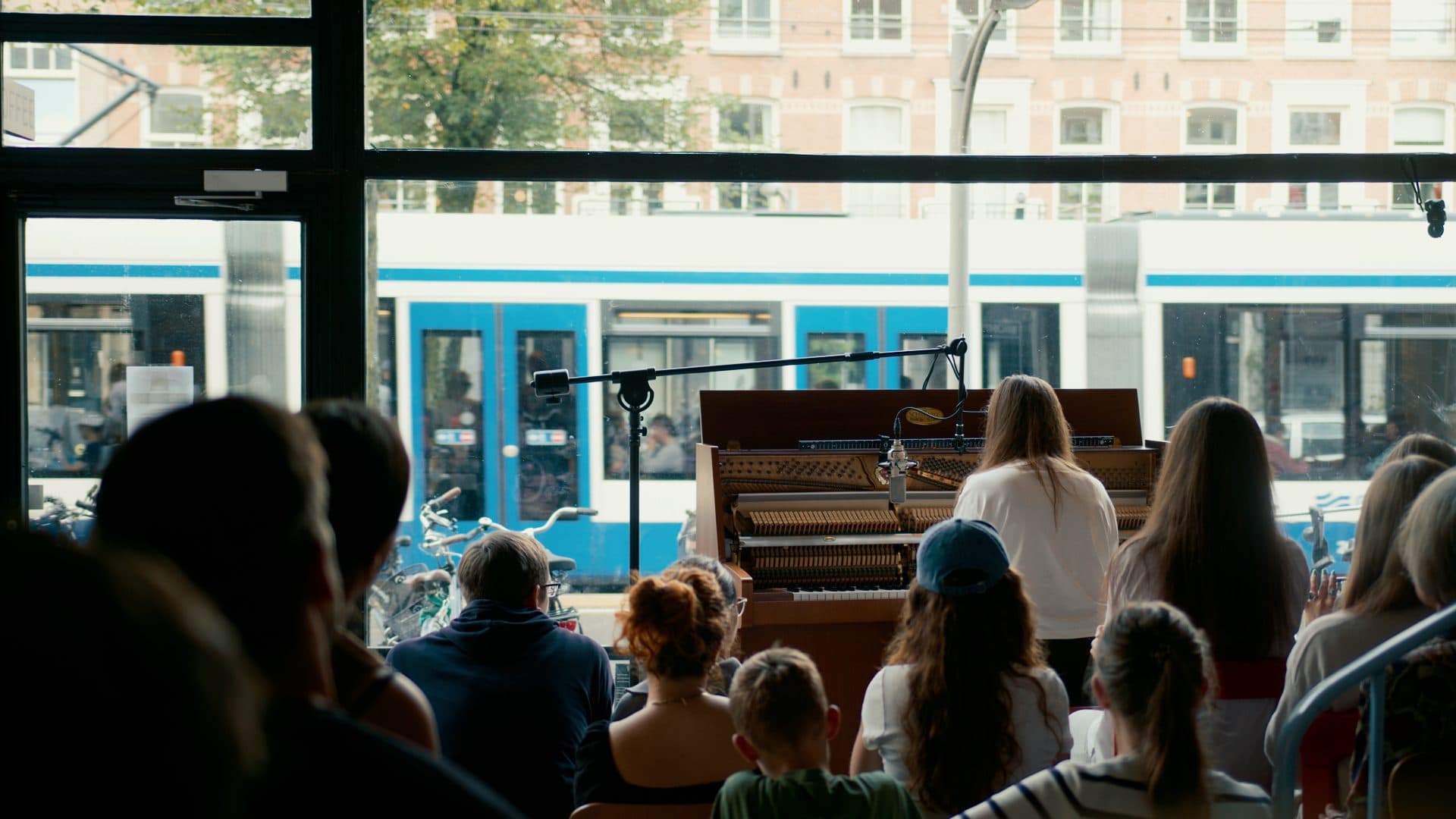 Muriel Bostdorp at an opened upright piano by the window at Bouncespace, the audience seated close, a blue and white tram passing outside