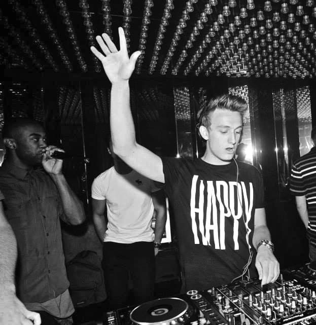 Ivo Adelaar as Vida behind the decks under the bulb ceiling of Jimmy Woo in Amsterdam, one arm raised, an MC on the microphone beside him