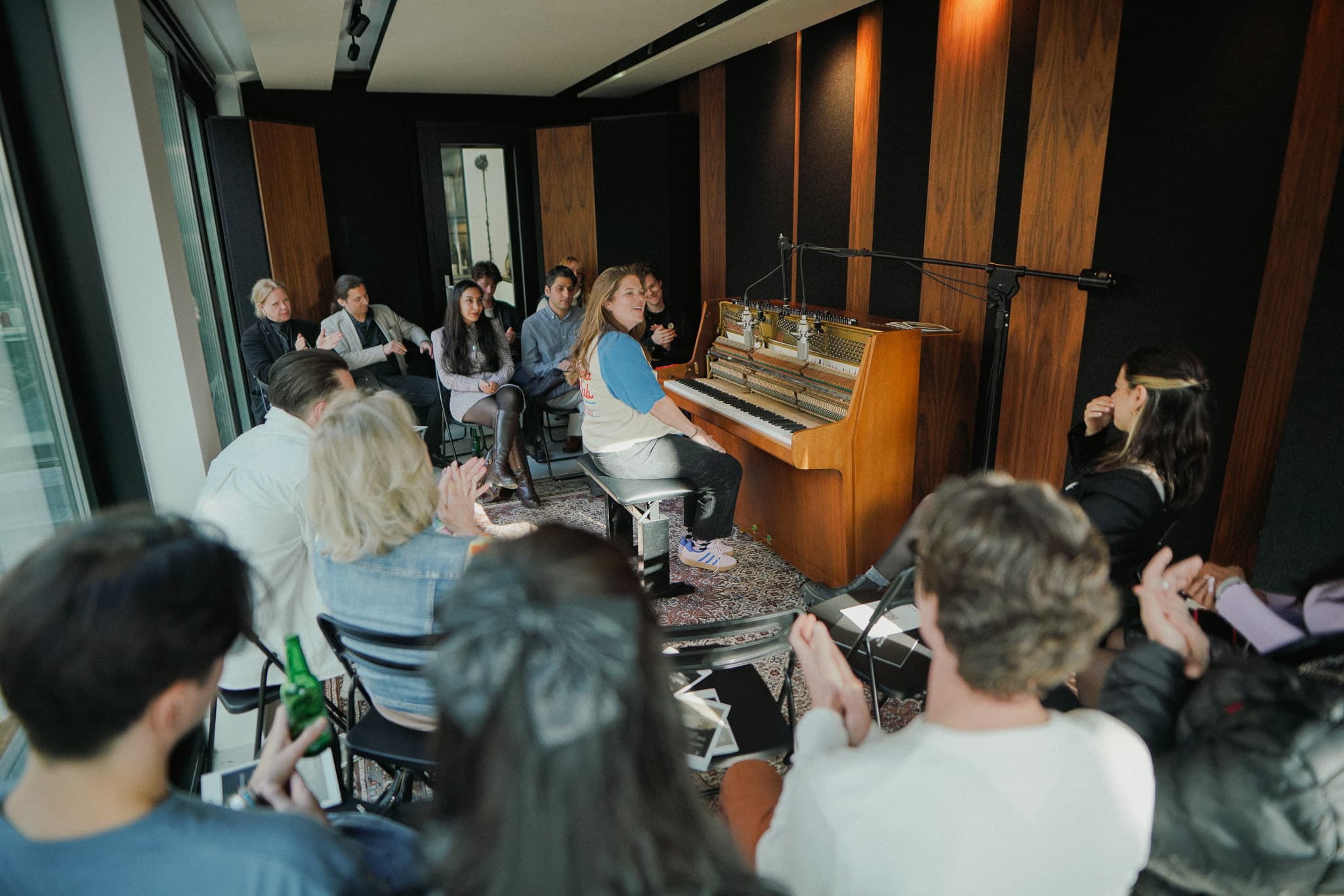 A pianist turning from the opened upright piano as the audience applauds, seated close around her in the studio